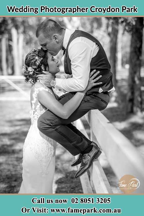 Photo of the Groom sitting on the fence while kissing the Bride on the forehead Croydon Park NSW 2133